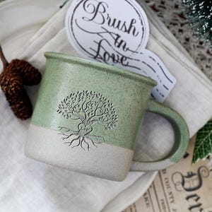 Peut inclure: Une tasse en céramique bicolore avec une partie supérieure vert sauge mouchetée et une base beige mate. La tasse présente un motif d'arbre de vie en relief avec un cœur au centre contenant les lettres L et V. Un autocollant indique Brush In Love.