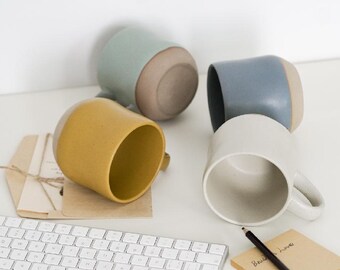 Könnte beinhalten: Vier Keramikbecher in Senfgelb, Hellblau, Graublau und Off-White. Die Becher haben ein zweifarbiges Design mit einem naturfarbenen Boden. Eine weiße Tastatur, ein Bleistift und ein Notizblock sind ebenfalls zu sehen.
