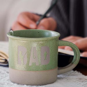 May include: Green ceramic mug with a white glaze and the word "DAD" written in a stylised font. The letters are outlined with a white border and include a fishing rod and fish graphic.