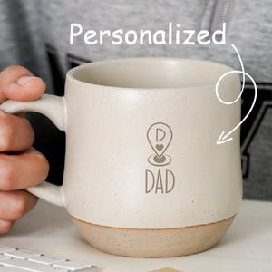 May include: Cream-colored ceramic mug with a speckled finish and a tan base. The mug features a location pin graphic with the word "DAD" below it. The word "Personalized" is written above the mug.
