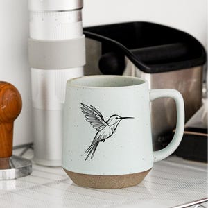 Puede incluir: Taza de café de cerámica azul claro con una ilustración de un colibrí. La taza tiene una base marrón y un asa grande. En el fondo se ven otros equipos para preparar café, como un molinillo y un recipiente.