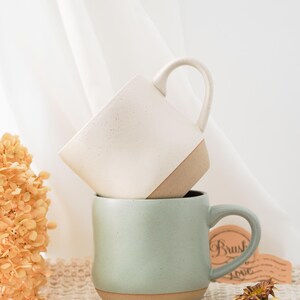 May include: Two ceramic mugs stacked on top of each other. The top mug is white with brown speckles and a brown rim. The bottom mug is a light green color with a brown rim. The mugs are sitting on a white crocheted surface.