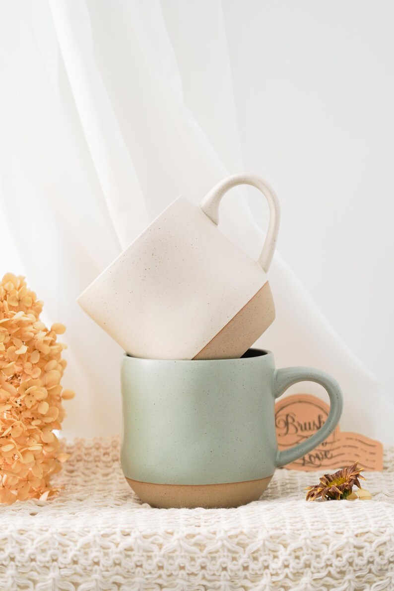 May include: Two ceramic mugs stacked on top of each other. The top mug is white with brown speckles and a brown rim. The bottom mug is a light green color with a brown rim. The mugs are sitting on a white crocheted surface.