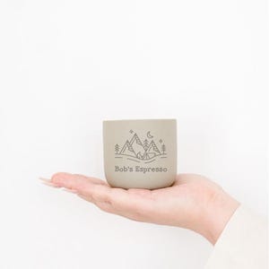 May include: A small, light grey espresso cup held in a hand. The cup has a mountain and moon design with the text "Bob's Espresso". The cup is cylindrical and appears to be made of ceramic.
