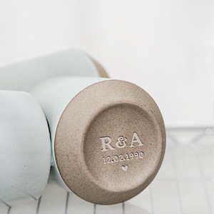May include: A light blue ceramic mug with a brown base, engraved with the initials "R&A", the date "12.02.1990", and a heart symbol. The mug is on a wire rack. The mug is likely for beverages.