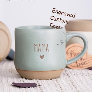 May include: A light green ceramic mug with a beige bottom. The mug has a handle and is engraved with the word "MAMA" and a small heart. The mug has a capacity of 17.5 ounces or 520 milliliters.