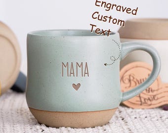 May include: A light green ceramic mug with a beige bottom. The mug has a handle and is engraved with the word "MAMA" and a small heart. The mug has a capacity of 17.5 ounces or 520 milliliters.