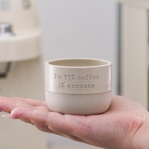 以下が含まれることがあります： 「I'm 99% coffee 1% excuses」の文字がエンボス加工された、オフホワイトのセラミックコーヒーカップ。ぼやけた背景の中で、人の手でカップが持たれています。