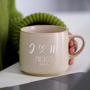 May include: A beige ceramic mug with a handle. The mug has the text "J & M ENGAGED 02.14.25" engraved on the front.