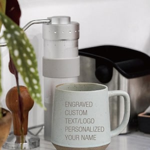 May include: A light blue ceramic mug with a brown base, featuring the text "ENGRAVED CUSTOM TEXT/LOGO PERSONALIZED YOUR NAME." A silver coffee grinder and other coffee-making equipment are in the background.