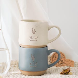 Könnte beinhalten: Zwei gestapelte Keramikbecher. Der obere Becher ist cremefarben mit gesprenkelter Oberfläche, der untere Becher blau. Beide Becher haben ein Blattdesign und die Initialen "L + J" mit dem Datum "06.03.2025" eingraviert. Rustikales, handgefertigtes Aussehen.