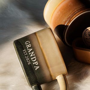May include: A ceramic mug with the word "GRANDPA" and "EST.2026" printed on the side. The mug has a handle and a two-tone color scheme of green and tan. Another mug is visible in the background, along with a wooden item.
