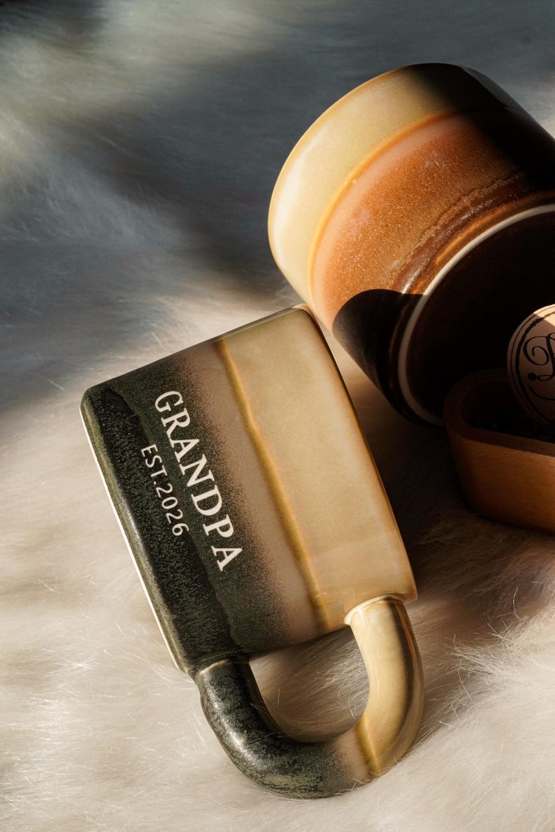 May include: A ceramic mug with the word "GRANDPA" and "EST.2026" printed on the side. The mug has a handle and a two-tone color scheme of green and tan. Another mug is visible in the background, along with a wooden item.