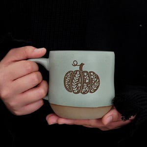 Può includere: Una tazza in ceramica azzurra con un disegno di zucca marrone. La zucca è decorata con motivi floreali e di foglie. Il fondo della tazza è di colore marrone chiaro. La tazza è tenuta con entrambe le mani.
