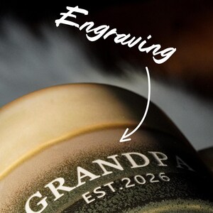 May include: Close-up of a ceramic mug with the word "GRANDPA" and "EST. 2026" in white lettering. The mug has a gradient color scheme, transitioning from a light tan to a dark green. The word "Engraving" is written above the mug.