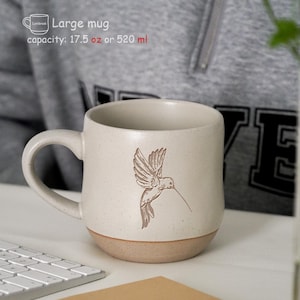 Puede incluir: Una taza grande de cerámica moteada con un diseño de colibrí. La taza es de color blanquecino con una base beige y una capacidad de 520 ml. La taza tiene un asa grande y está sobre una superficie blanca.