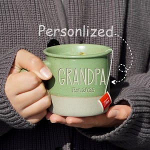May include: Green ceramic mug with the word "GRANDPA" and "Est. 2025" in white lettering. The mug is being held by a person wearing a gray sweater.