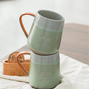Puede incluir: Dos tazas de cerámica apiladas con base verde claro, borde gris y asa naranja. Cada taza tiene el texto "Lucy & Jack" y un símbolo de corazón. Un portavasos de madera es visible en el fondo.