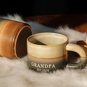 May include: A ceramic mug with a gradient design, transitioning from beige to olive green. The word "GRANDPA" and the year "EST.2026" are printed in white. Another mug is visible, lying on its side. The mugs are on a white, fluffy surface.