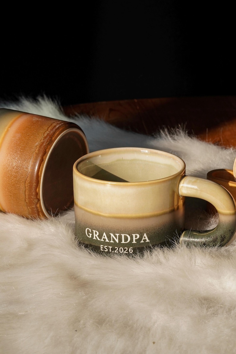 May include: A ceramic mug with a gradient design, transitioning from beige to olive green. The word "GRANDPA" and the year "EST.2026" are printed in white. Another mug is visible, lying on its side. The mugs are on a white, fluffy surface.