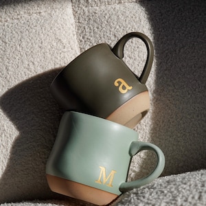 Pode incluir: Duas canecas de cerâmica, uma verde-oliva e outra verde-azulado claro, empilhadas. A caneca verde-oliva tem a letra dourada "a", e a caneca verde-azulado tem a letra dourada "M". Ambas as canecas têm uma base bege e uma alça curva.