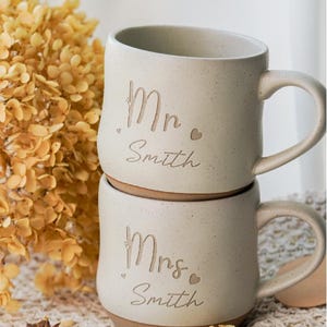 May include: Two white ceramic mugs with brown accents. The top mug says "Mr. Smith" and the bottom mug says "Mrs. Smith".