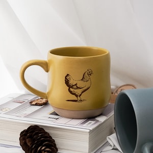 May include: Mustard yellow ceramic mug with a brown illustration of a hen. The mug has a rounded shape and a large handle. The bottom of the mug is a light beige color. The mug is sitting on a book.