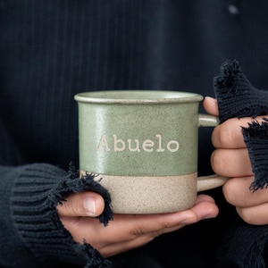 May include: A two-tone ceramic mug with the word "Abuelo" in a light green color. The mug is held by a person wearing a black sweater with distressed details.