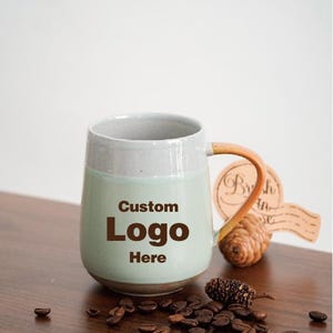 May include: A ceramic mug with a light green body and a gray rim. The handle is a light brown color. The mug has the words "Custom Logo Here" printed on the front. Coffee beans and a small pinecone are scattered around the mug.