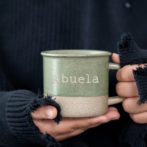 以下が含まれることがあります： 「Abuela」の文字が入った、明るい色のツートーンセラミックマグ。マグの上半分はくすんだグリーン、下半分はまだらなベージュです。黒いセーターを着た人が、マグカップを持っています。