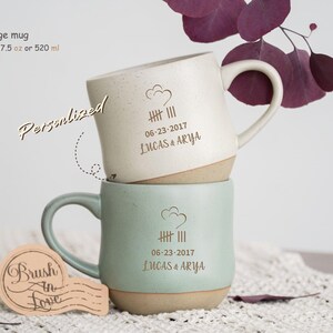 Puede incluir: Dos tazas de cerámica apiladas, una color crema y otra verde menta, con base beige. Cada taza tiene un asa y está personalizada con una fecha y nombres. La taza crema tiene un diseño de nube. Capacidad: 520 ml.