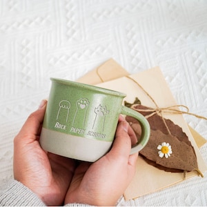 Puede incluir: Taza de cerámica verde con un borde blanco. La taza tiene un diseño de huella de pata y el texto "Piedra Papel Tijeras".