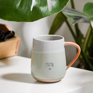Puede incluir: Una taza de cerámica con cuerpo verde claro, borde gris y asa color melocotón. La palabra "MAMA" y un corazón están impresos en la parte delantera. La taza está sobre una superficie blanca, con una gran hoja verde al fondo.