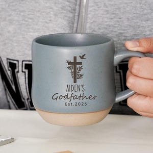 Può includere: Una tazza in ceramica blu con base beige. La tazza presenta un disegno a croce e colomba con la scritta "AIDEN'S Godfather Est. 2025". La tazza ha un manico ed è tenuta da una persona.