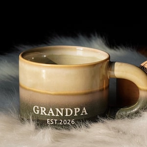 May include: A ceramic mug with a two-tone design, featuring a light beige top and a dark green base. The mug has a curved handle and the word "GRANDPA" and "EST. 2026" printed in white. The mug is resting on a white, fluffy surface.