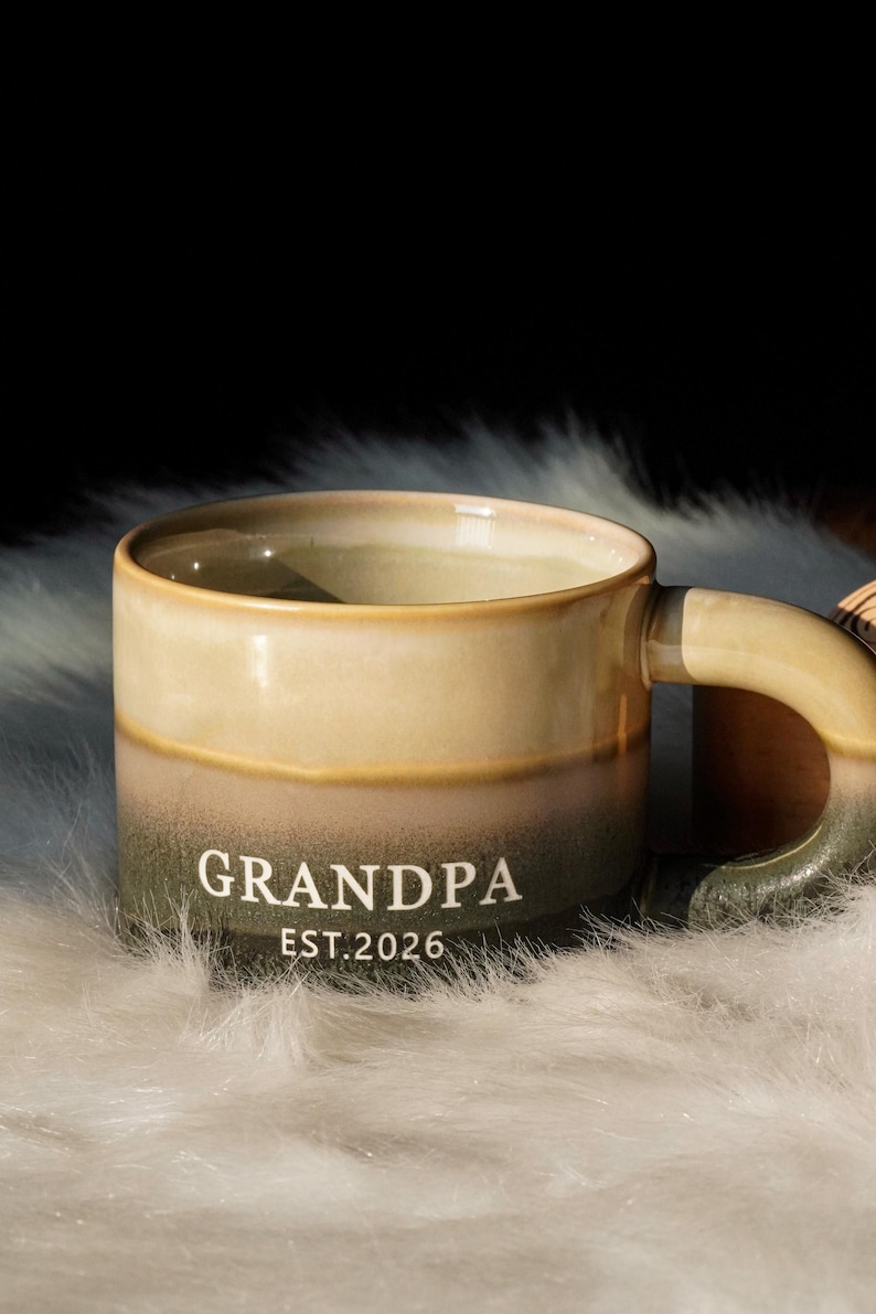 May include: A ceramic mug with a two-tone design, featuring a light beige top and a dark green base. The mug has a curved handle and the word "GRANDPA" and "EST. 2026" printed in white. The mug is resting on a white, fluffy surface.