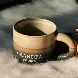 May include: A ceramic mug with a gradient color scheme, transitioning from beige to dark green. The word "GRANDPA" and the year "EST.2026" are printed in white. The mug has a large handle and sits on a fluffy, white surface.