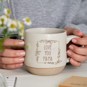 以下が含まれることがあります： クリーム色のセラミックマグカップ。底はベージュ色で、「LOVE YOU MAMA」の文字が花のフレームの中に描かれています。マグカップには取っ手があり、人が持っています。