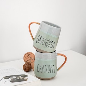 May include: Two ceramic mugs, one stacked on the other, with the words "GRANDMA" and "GRANDPA" in black script. The mugs have a light green band, gray top, and orange handle. A pinecone and a book are in the foreground.