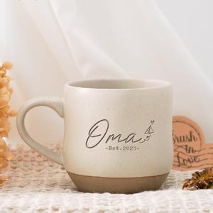 May include: A large white ceramic mug with a brown bottom. The mug has the word "Oma" written on it in cursive with a paper airplane and heart above it. The text "Est. 2025" is written below the word "Oma".