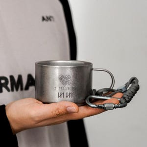 May include: A small, brushed metal mug with a handle and the text "I Still Do" and a heart design. A grey carabiner clip is attached to the mug. The mug is held in a person's hand.