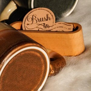 May include: Close-up of a brown ceramic mug with a white rim and handle. A wooden holder with the text "Brush In" is in the background. The mug is on a white, fluffy surface. Other mugs are visible.