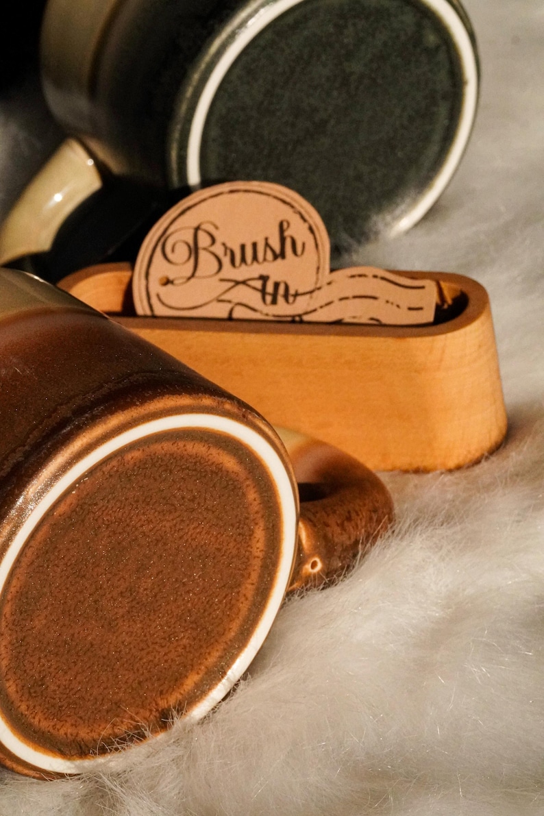 May include: Close-up of a brown ceramic mug with a white rim and handle. A wooden holder with the text "Brush In" is in the background. The mug is on a white, fluffy surface. Other mugs are visible.