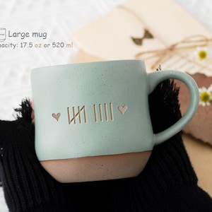 Puede incluir: Una taza de cerámica verde claro con un borde marrón y un asa. La taza tiene un diseño de corazón y números romanos. La taza tiene una capacidad de 520 ml.
