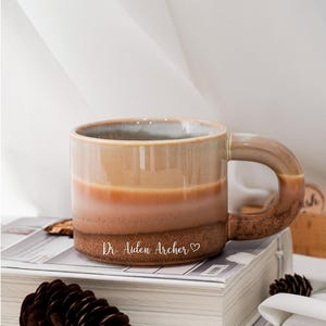 Puede incluir: Una taza de cerámica blanca con un asa negra y el texto "But First, Coffee" en negro, con una fuente de estilo manuscrito. La taza está sobre una superficie blanca.