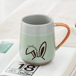 Puede incluir: Una taza de cerámica con cuerpo verde claro, borde gris y asa marrón. La taza presenta un diseño de conejito negro y el nombre "Anderson". La taza está sobre un libro abierto.