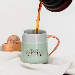 May include: A ceramic mug with a light green base and gray top is being filled with dark coffee. The mug features a copper-colored handle and a design of two elephants with intertwined trunks. A wooden coaster with the word "Brush" is in the background.
