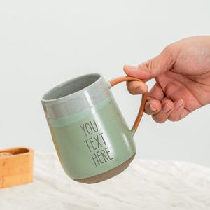 Puede incluir: Una taza de cerámica con cuerpo verde claro, borde gris y asa marrón. La taza tiene el texto "YOU TEXT HERE" impreso en la parte delantera. La taza está sujeta por una mano.