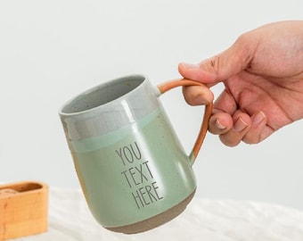 Op de afbeelding: Een keramische mok met een lichtgroene body, grijze rand en bruin handvat. De mok heeft de tekst "YOU TEXT HERE" op de voorkant gedrukt. De mok wordt door een hand vastgehouden.