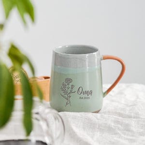 Puede incluir: Una taza de cerámica con cuerpo verde claro, borde gris y asa naranja. La taza presenta un diseño floral y la palabra "Oma" con "Est.2025" impresa debajo. La taza está sobre una tela blanca.